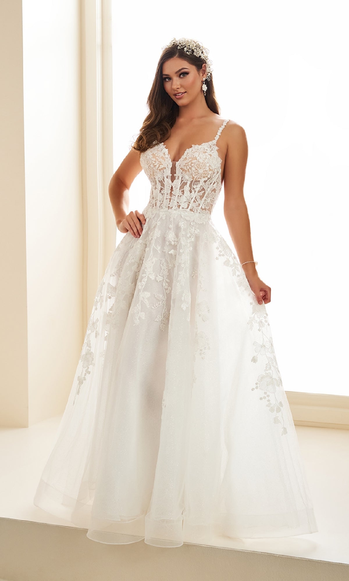 A woman stands indoors by a sunlit window wearing the Ellie Wilde EW36015, a sleeveless white long prom dress with a corset bodice, lace embroidery, floral appliqués, and a full tulle skirt.